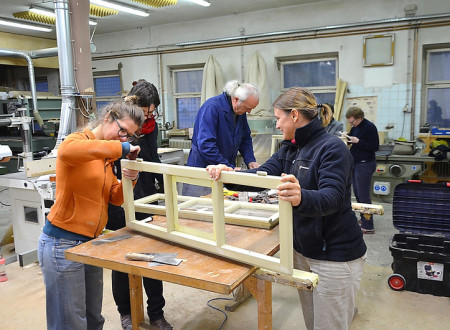 Ženski v delavnici, v kateri so še štiri osebe, obnavljata lesen okvir oken s tremi stekli