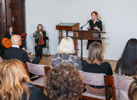 Ministrica za kulturo stoji za govornim pultom z mikrofonom, na njeni desni sedita violinistka in kitaristka, pred njo na stolih sedijo ljudje