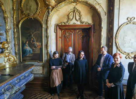 Soba v Gruberjevi palači z okvirjenimi slikami na steni in obokanimi vrtati, nad njimi in na njih ornamenti, pred njimi stoji ministrica v družbi s petimi udeleženci odprtja