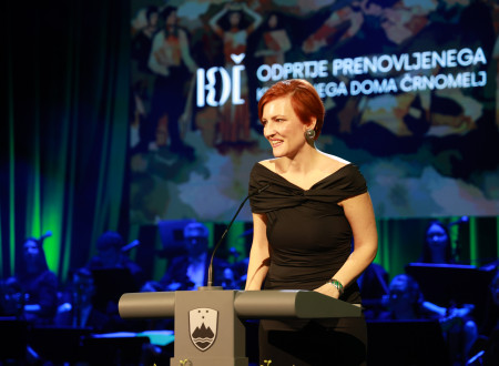 Ministrica za kulturo stoji za govornim pultom z mikrofonom, za njo napis na zaslonu Odprtje prenovljenega Kulturnega doma Črnomelj 