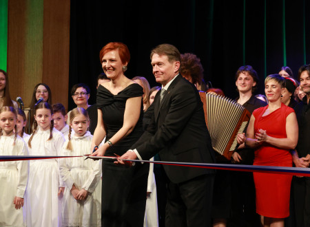 Nasmejana ministrica za kulturo in župan na odru režeta otvoritveni trak, za njima nastopajoči v okviru kulturnega programa ob odprtju kulturnega doma