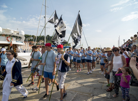 Obmorska parada, v kateri skupina ljudi v mornarskih in piratskih kostumih koraka ob nabrežju, nekateri nosijo črne zastave s simbolom lobanje. Ob poti stojijo gledalci, v ozadju pa so vidni privezi z jadrnicami.