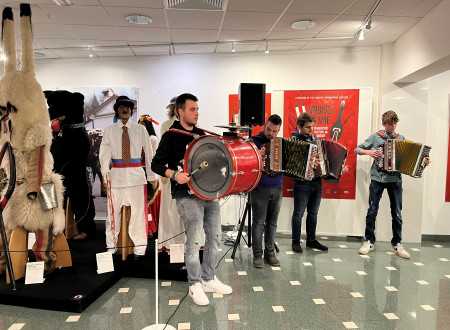 Trije harmonikaši in en bobnar igrajo pred razstavljenimi šemami, med njimi svetli in črni kurent