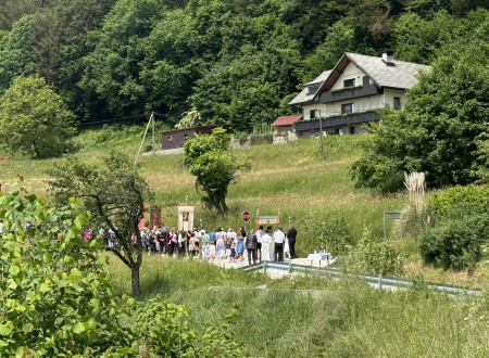 Ljudje v procesiji na vaški cesti sredi hribovite zelene pokrajine, nad cesto hiša ob gozdu