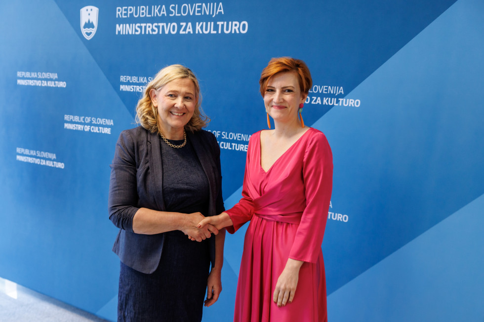 Nova in dosedanja ministrica se rokujeta pred modrim platnom z logotipom Ministrstva za kulturo