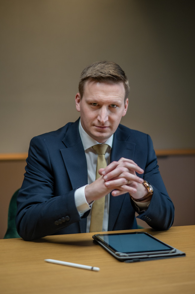 Moški s svetlo rjavimi in kratko pristriženimi lasmi, v modri obleki s sklenjenimi dlanmi in s komolci naslonjenimi na mizo 