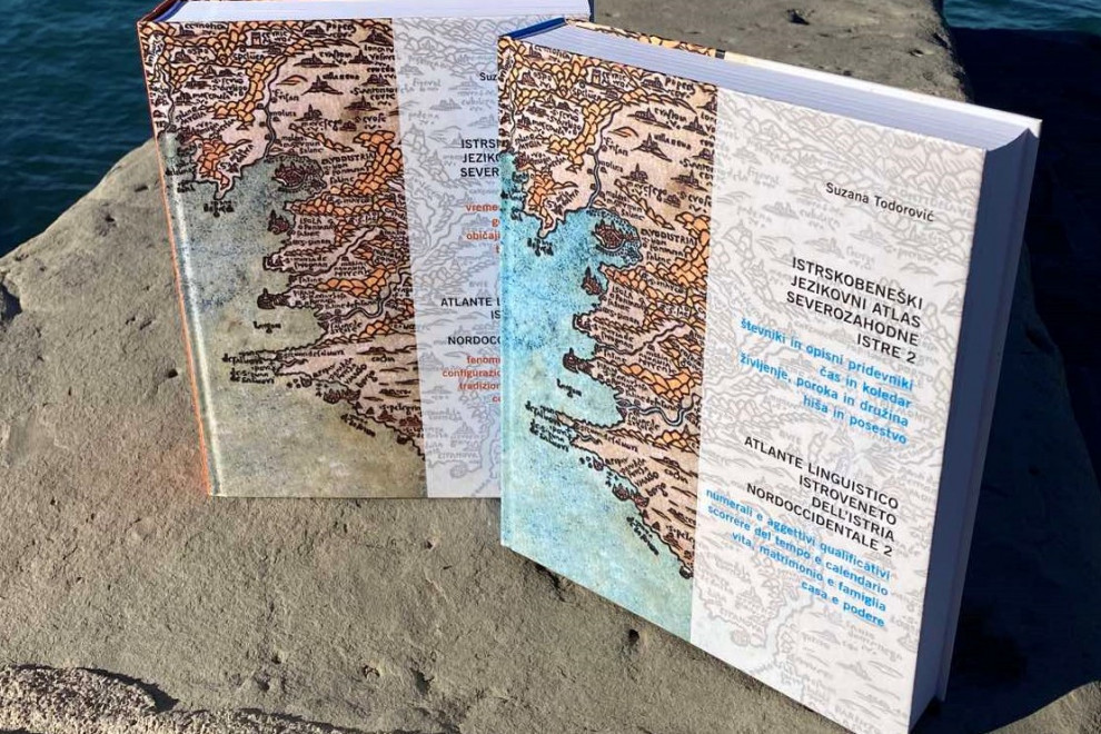 Naslovnici dveh zvezkov Istrskobeneškega jezikovnega atlasa severozahodne Istre, fotografirani na morskem pomolu.