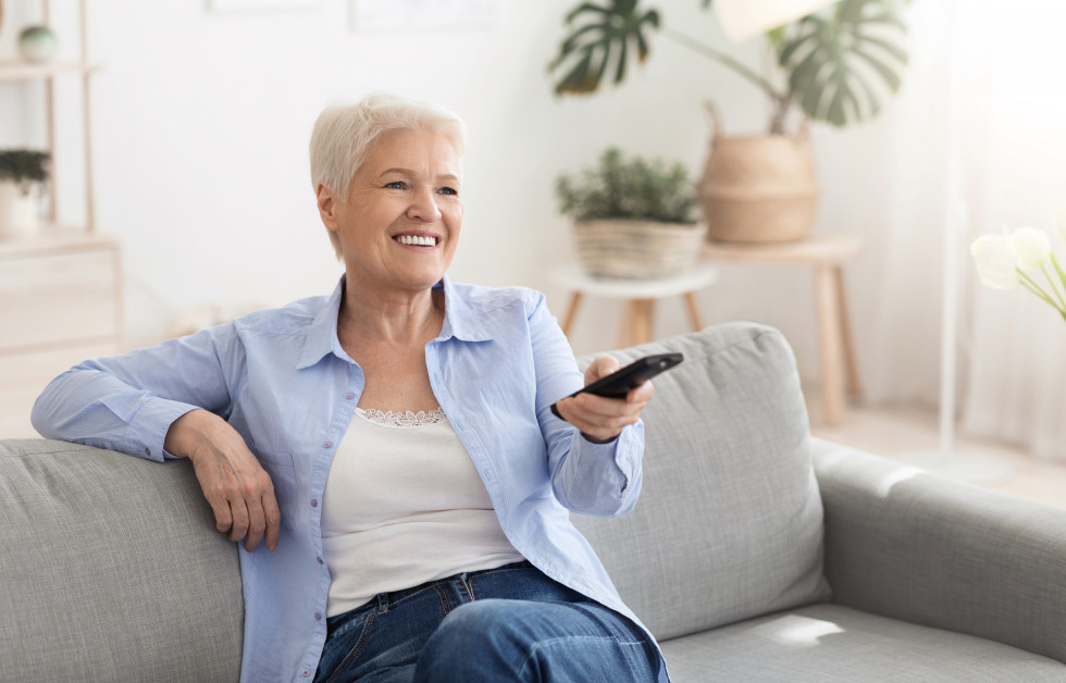 Smejoča se starejša gospa, ki spremlja TV program  z daljinskim upravljalcem v roki, na kavču v domačem okolju.