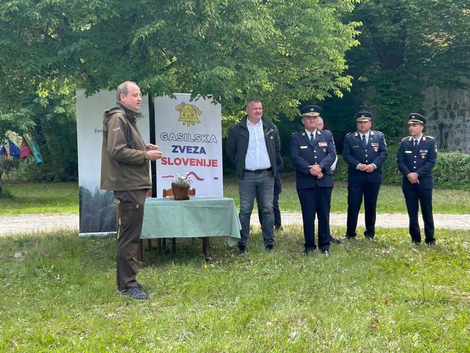 Govorec stoji na travi. Nekoliko stran moški v uniformah,  
