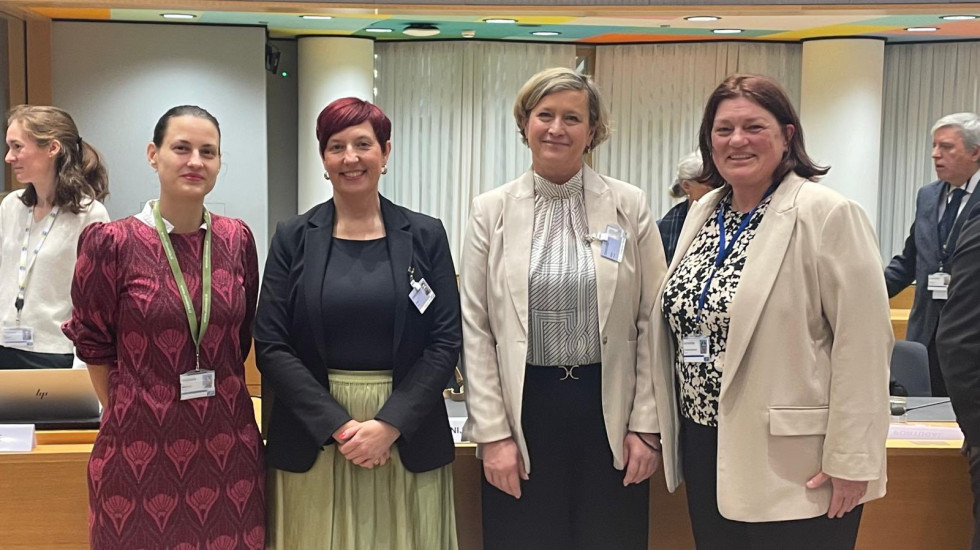 V sejni dvorani stoji državna sekretarka in tri predstavnice Slovenije obrnjene proti kameri, za njimi mize in okno