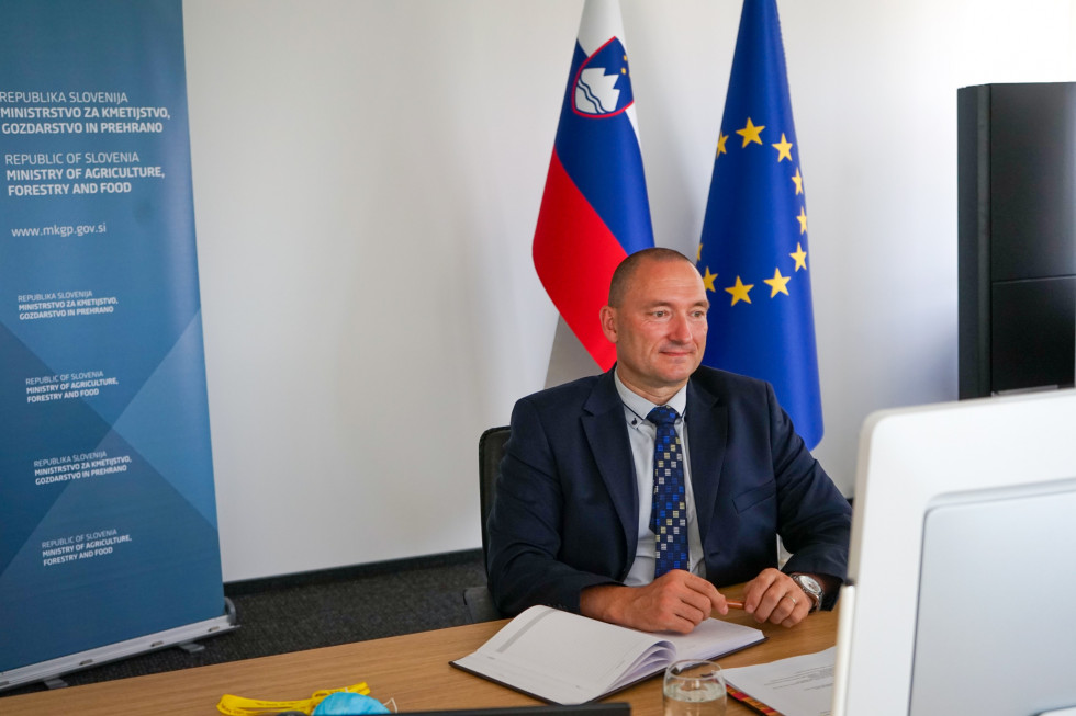 Minister  v obleki sedi pred računalnikom in spremlja dogajanje na ekranu pred njim. V ozadju slovenska zastava in zastava evropske unije