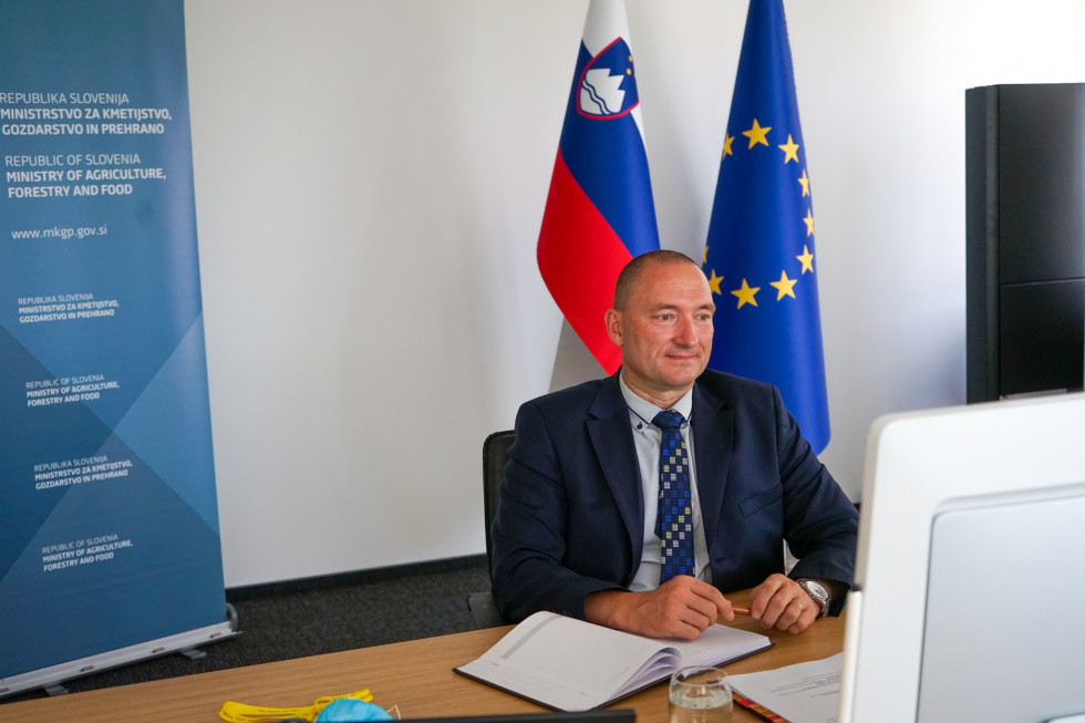 Minister v obleki sedi pred računalnikom in spremlja dogajanje na ekranu pred njim. V ozadju slovenska zastava in zastava evropske unije