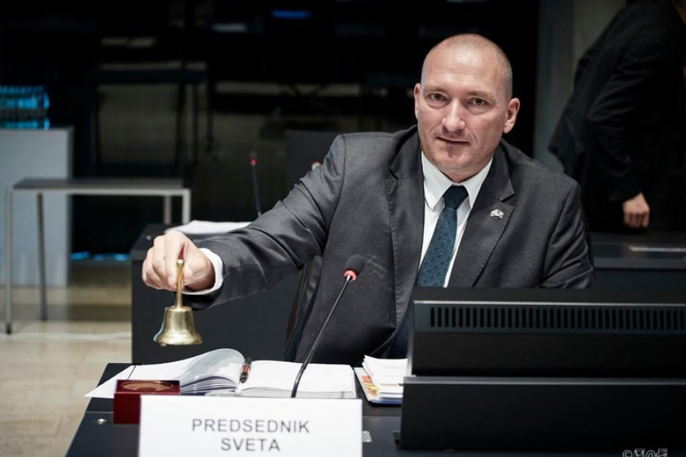 Minister ob začetku sveta. V rokah ima zvonček s katerim pozvoni za začetek zasedanja. 