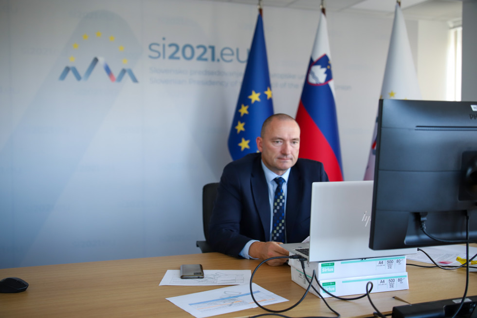 Minister sedi za mizo in spremlja dogajanje na ekranu. V ozadju zastave slovenskega predsedovanja, Slovenije in Evropske unije