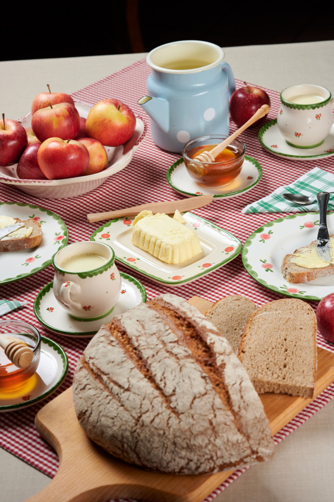 Na mizi so hleb kruha, rezina kruha, maslo, mleko v skodelici, med in jabolka v košari.