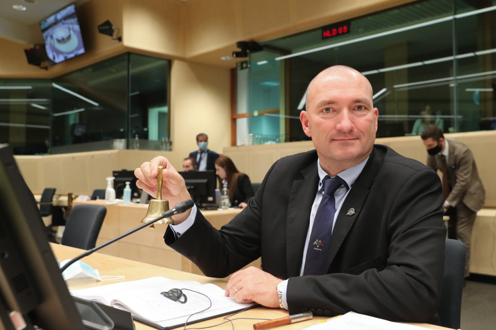 Minister sedi za mizo. V rokah ima zvonček s katerim oznanja začetek zasedanja.