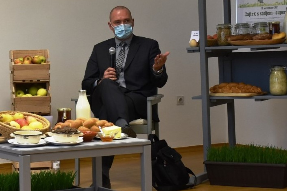 Moški v temno sivi obleki sedi z masko na obrazu govori v mikrofon v roki . Pred njim je nizka mizica s hrano, ob njem na desno se vidi stojalo.