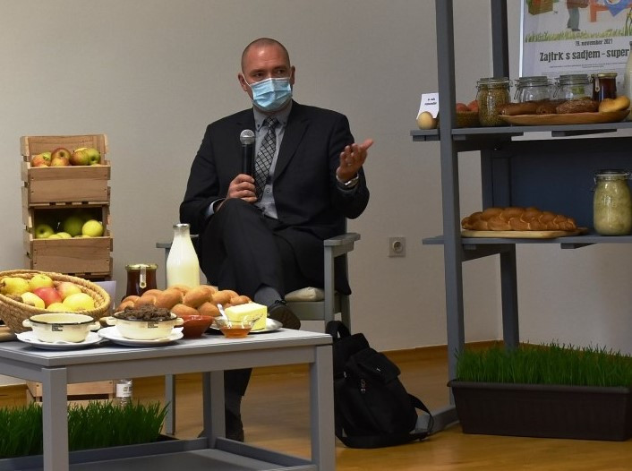 Moški v temno sivi obleki sedi z masko na obrazu govori v mikrofon v roki . Pred njim je nizka mizica s hrano, ob njem na desno se vidi stojalo.