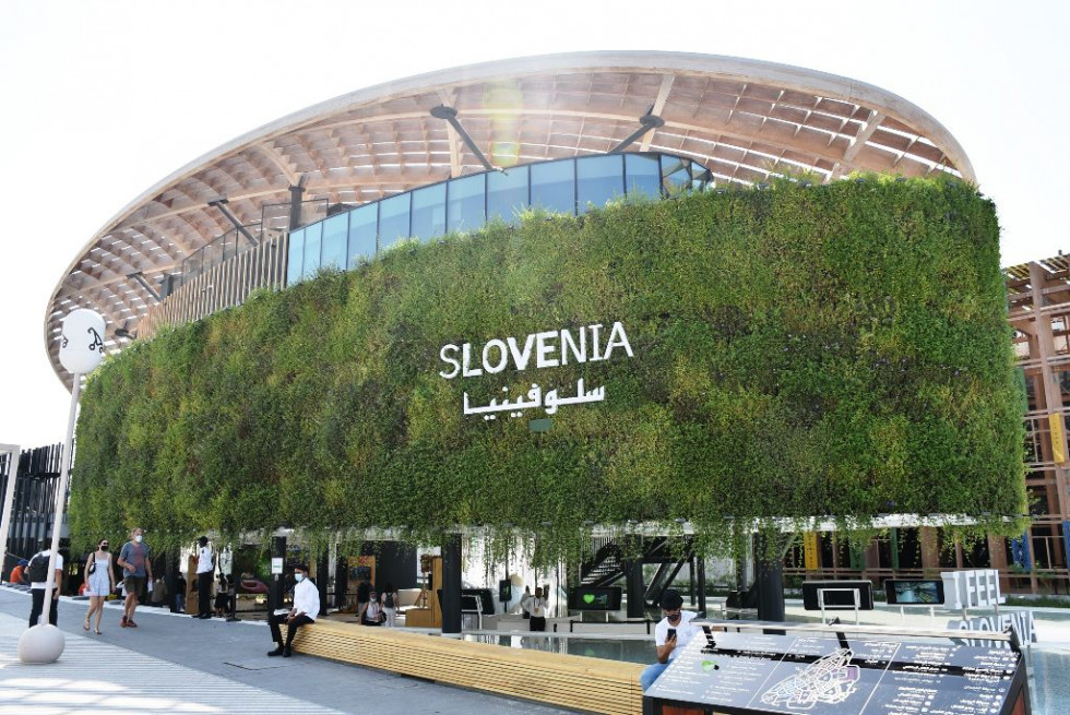 Slika slovenskega paviljona na razstavi EXPO. Pred njim klopca na kateri sedijo obiskovalci. 