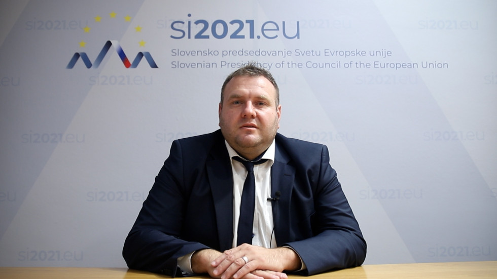Državni sekretar Irgolič sedi na stolu in spremlja zasedanje. Za njim napis slovenskega predsedovanja. 