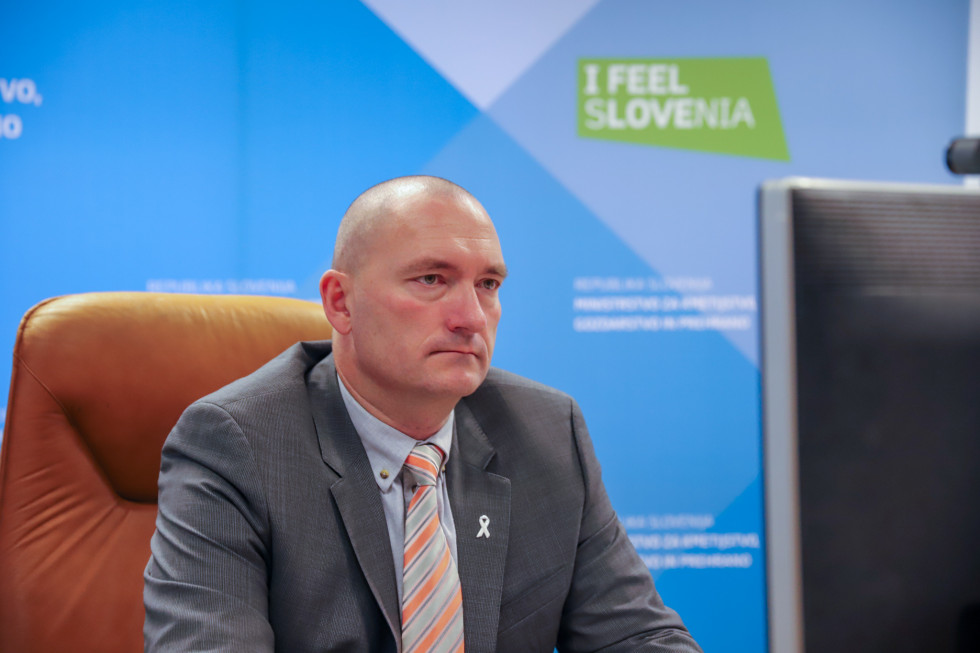 Minister sedi za mizo in spremlja dogodek. Za njim napis ministrstva in I feel Slovenia.