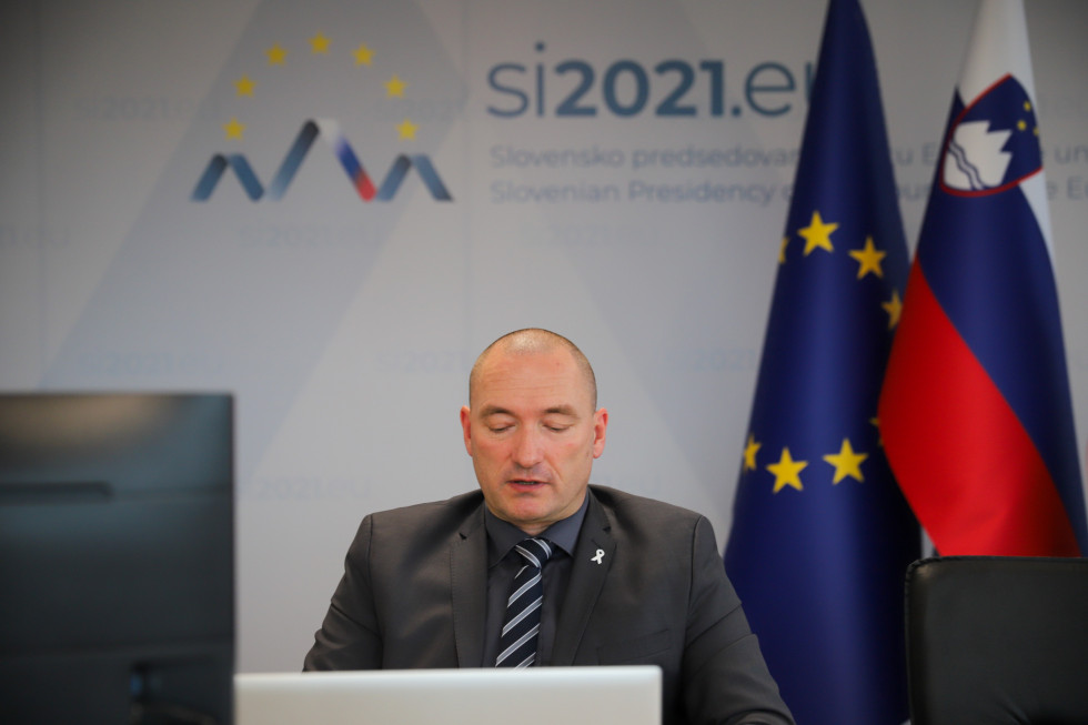 Minister dr. Jože Podgoršek spremlja dogajanje na ekranu. Za njim logotip predsedovanja Svetu Evropske Unije. 