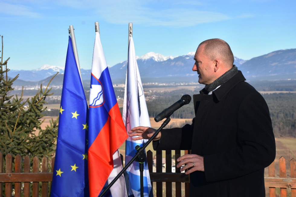 Minister govori v mikrofon. Ob njem so postavljene zastave, za njim pa se vidi gore. 