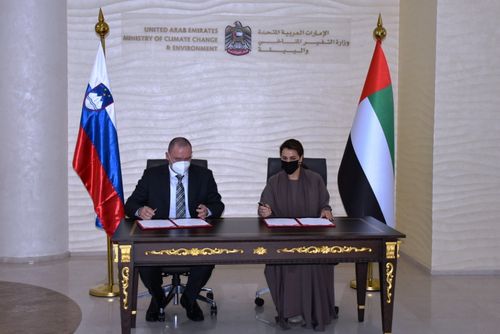 Slovenian minister signing the memorandum with  UAE Minister of Climate Change and Environment, Ms Mariam bint Mohammed Saeed Hareb Almheiri. Behind them the flags of both states. 