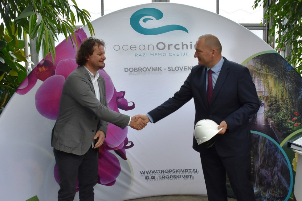 Minister in direktor podjetja se rokujeta. Za njima pano podjetja Ocean Orchids. 