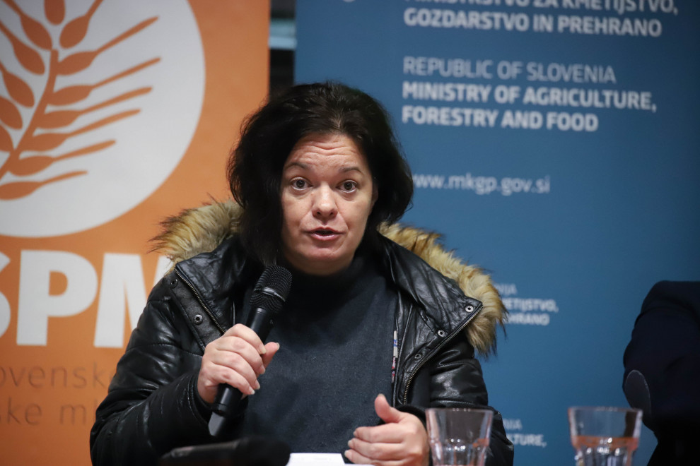 Državna sekretarka sedi za mizo in drži mikrofon v rokah. 