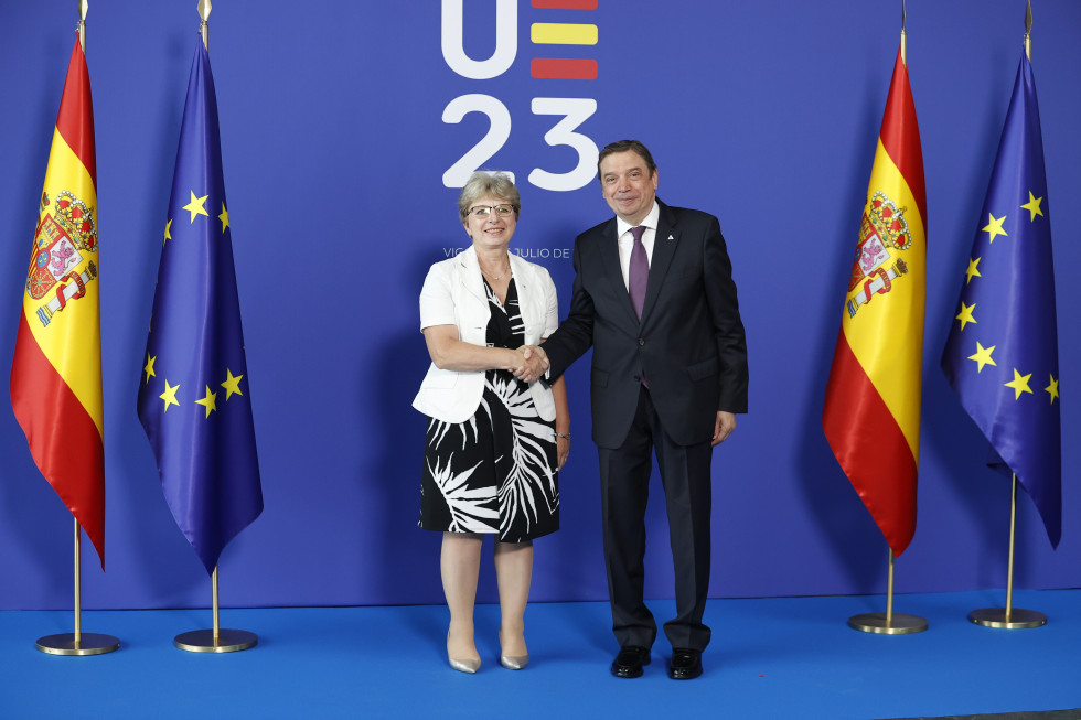 Ministrica in španski minister stojita pred zastavo in se rokujeta. 