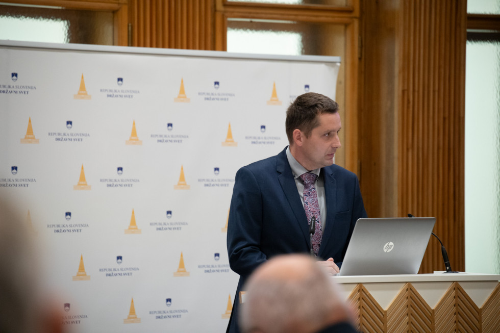 Državni sekretar stoji za govorniškim pultom in govori.