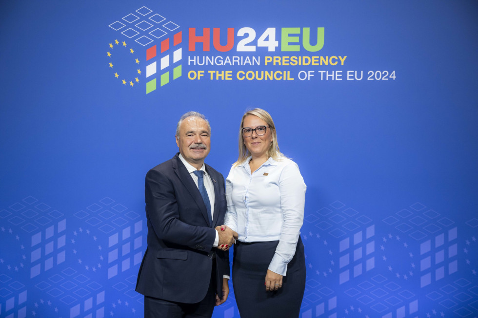Ministra stojita in se rokujeta pred modrim panojem s simbolom madžarskega predsedovanja Svetu EU in napisom Madžarsko predsedovanje Svetu EU 2024 v angleščini
