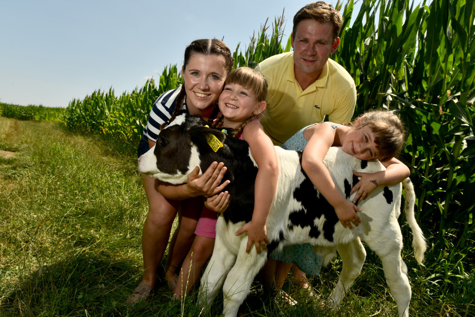 Mladi kmetje. Moški, ženska in dve deklici objemajo telička. Za njimi koruzno polje.