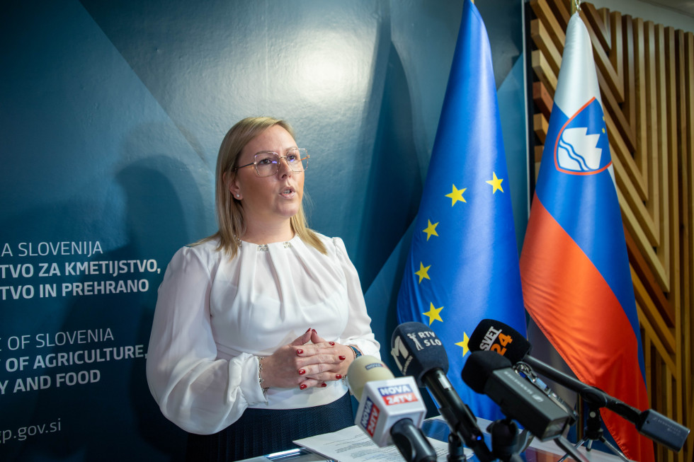 Ministrica stoji za govorniškim pultom in govori. 