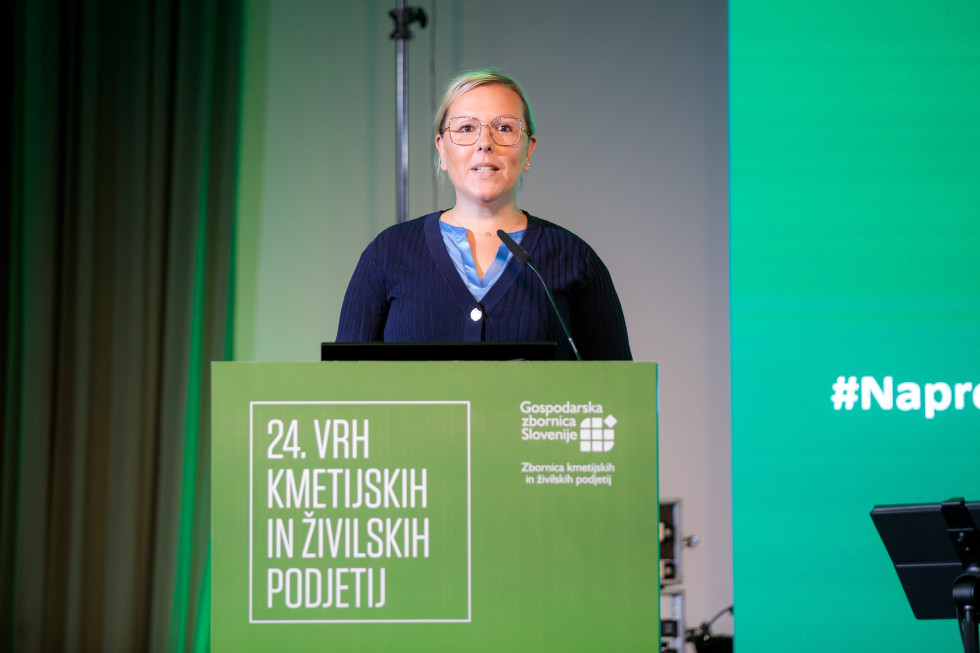 Ministrica stoji za govorniškim pultom in govori. 