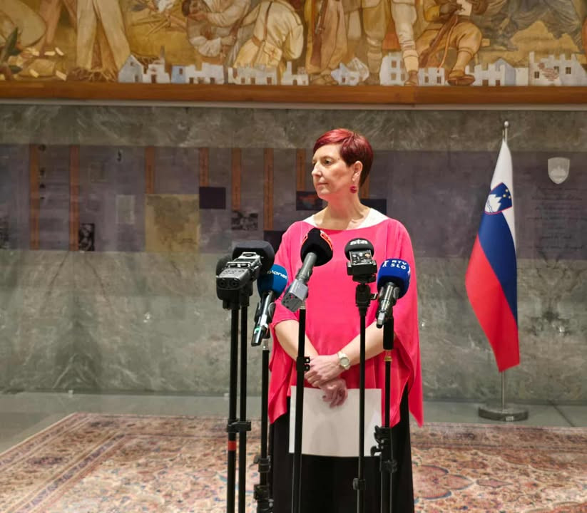 Državna sekretarka stoji pred mikrofoni in posluša. 