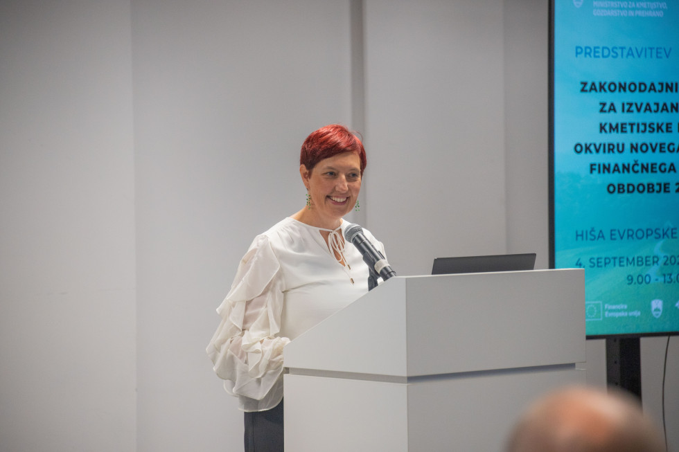 Državna sekretarka stoji za govorniškim pultom in govori.
