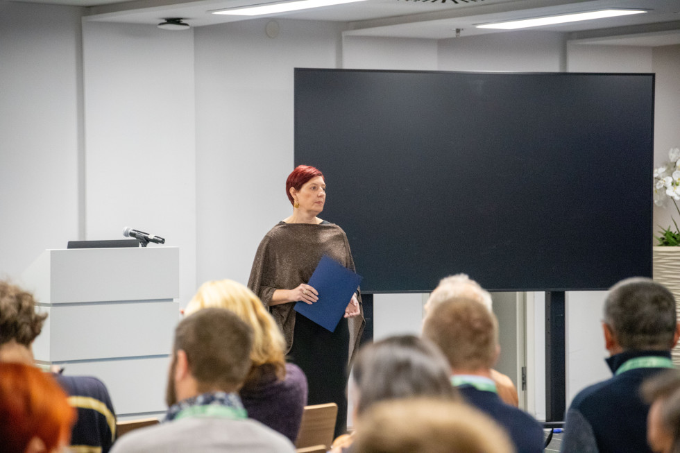 Oseba ženskega spola stoji in govori. Za njo je televizija. 