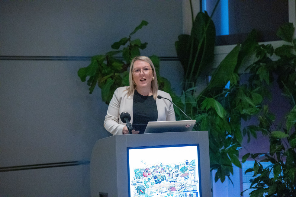 Oseba ženskega spola stoji za govorniškim pultom in govori. Za njo različne rože. 