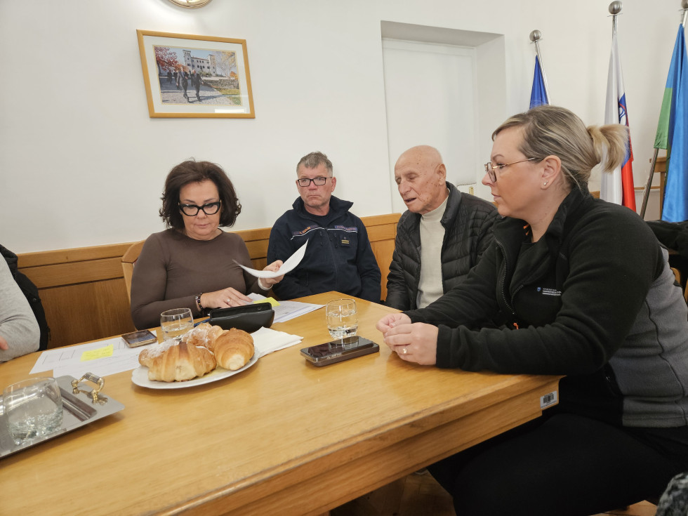 Osebe sedijo za mizo v dvorani in se pogovarjajo. 