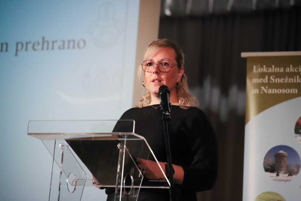 Oseba stoji za govorniškim pultom in govori. 