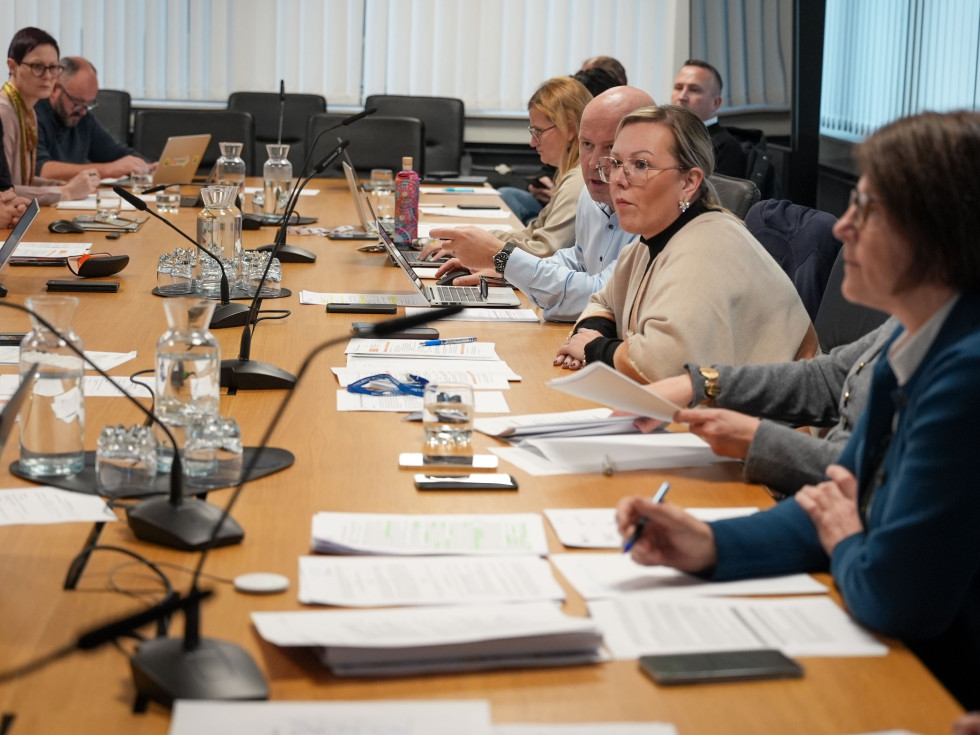 Več oseb sedi za leseno mizo. Pred njimi so papirji. Osebe se pogovarjajo.