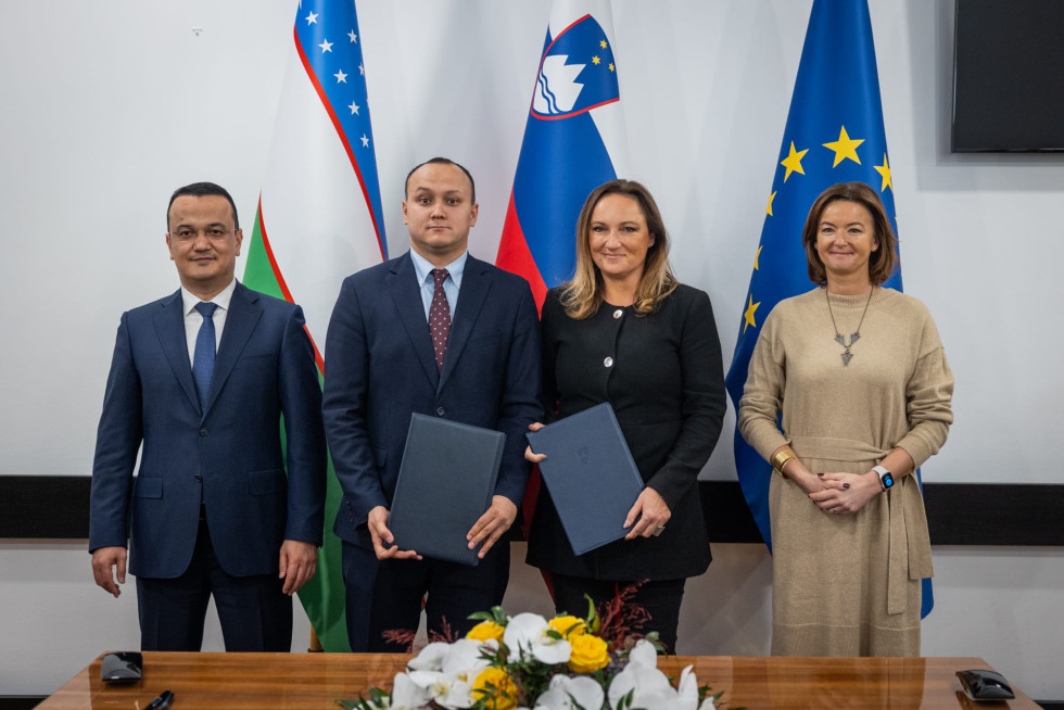 Več oseb stoji in se fotografira. Za njimi slovenska in uzbekistanska zastava. Pred njimi miza. 