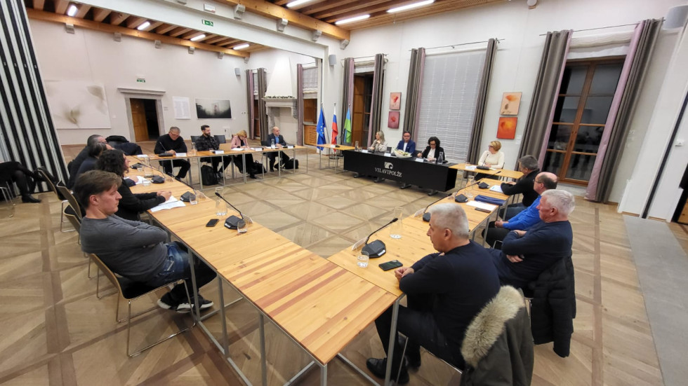 Več oseb sedi za mizo v dvorani. Med seboj se pogovarjajo. 