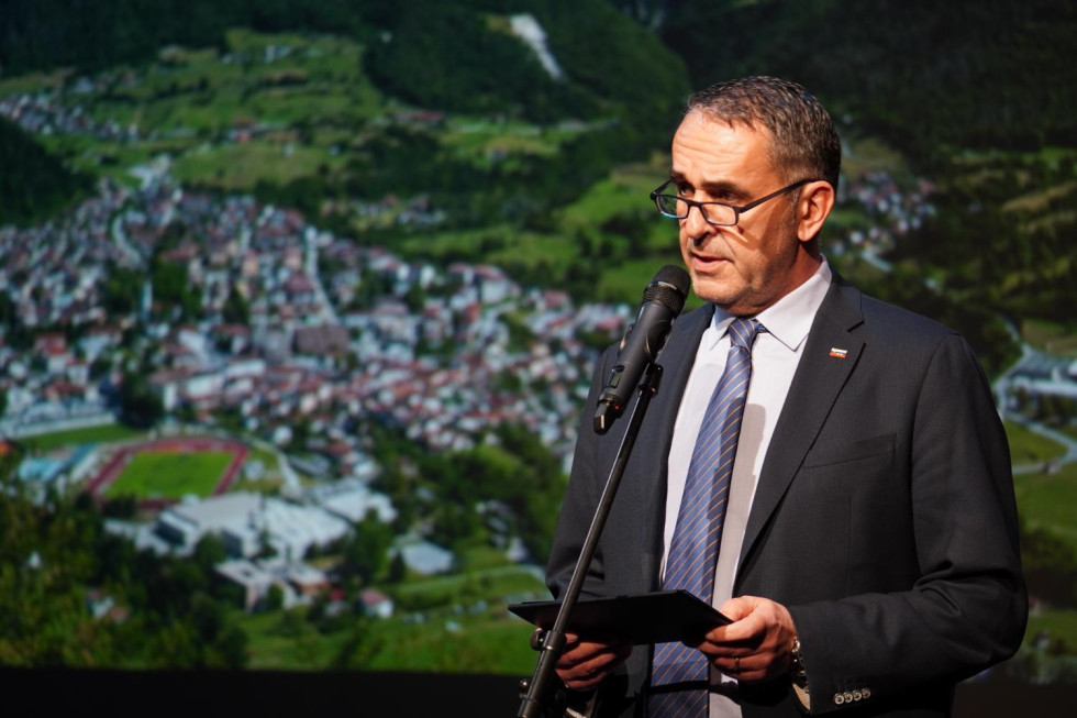 Oseba stoji za mikrofonom. V rokah drži govor. Na platnu za njim je prikazano mesto. 