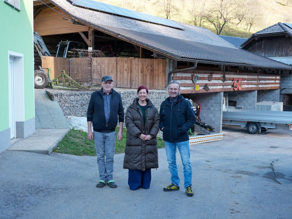 Tri osebe stojijo pred hlevom in se fotografirajo. 