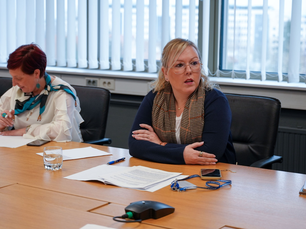 Ministrica Čalušić sedi za dolgo mizo, v ozadju okno