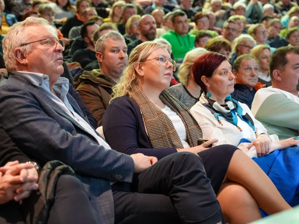 Več oseb sedi v dvorani in spremlja razpravo. 