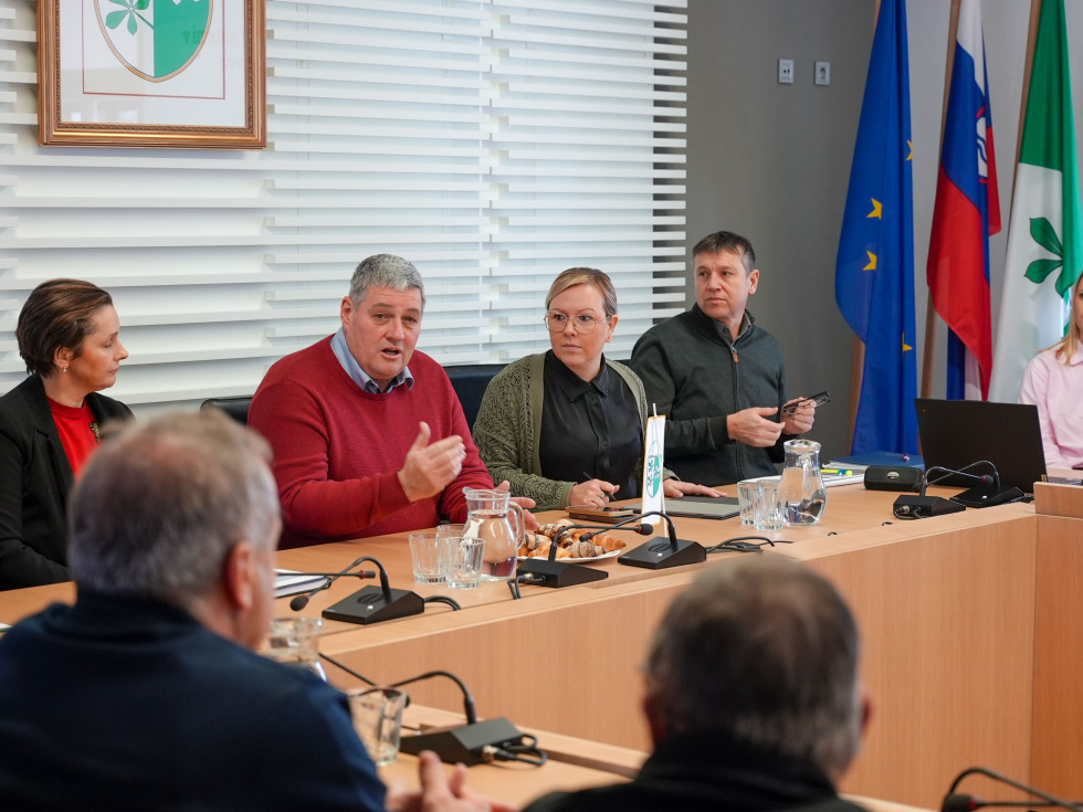 Več oseb sedi za mizo v dvorani in se pogovarjajo. 